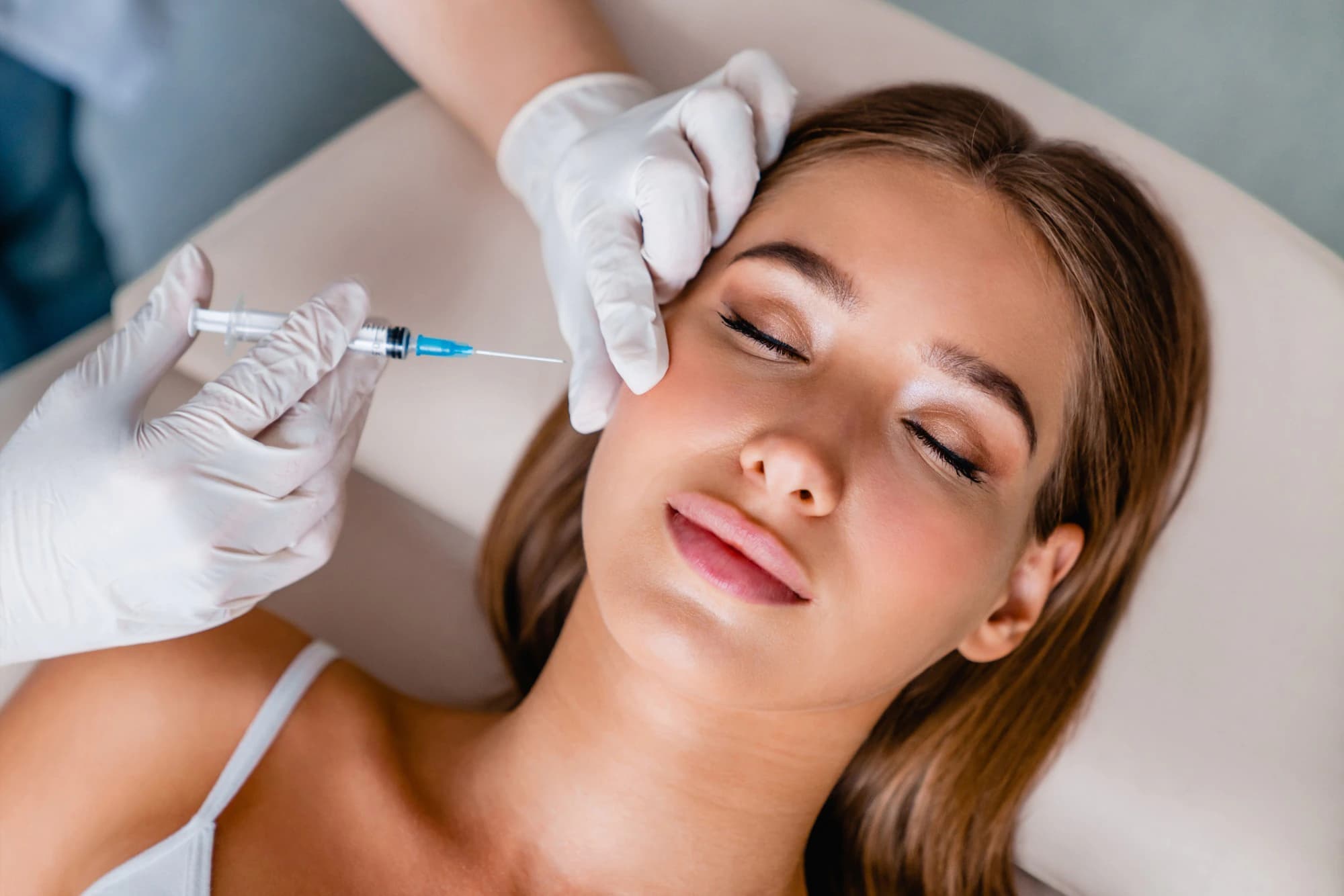 A woman lying back with her eyes closed while a gloved provider holds a syringe near her cheek during a Botox® treatment